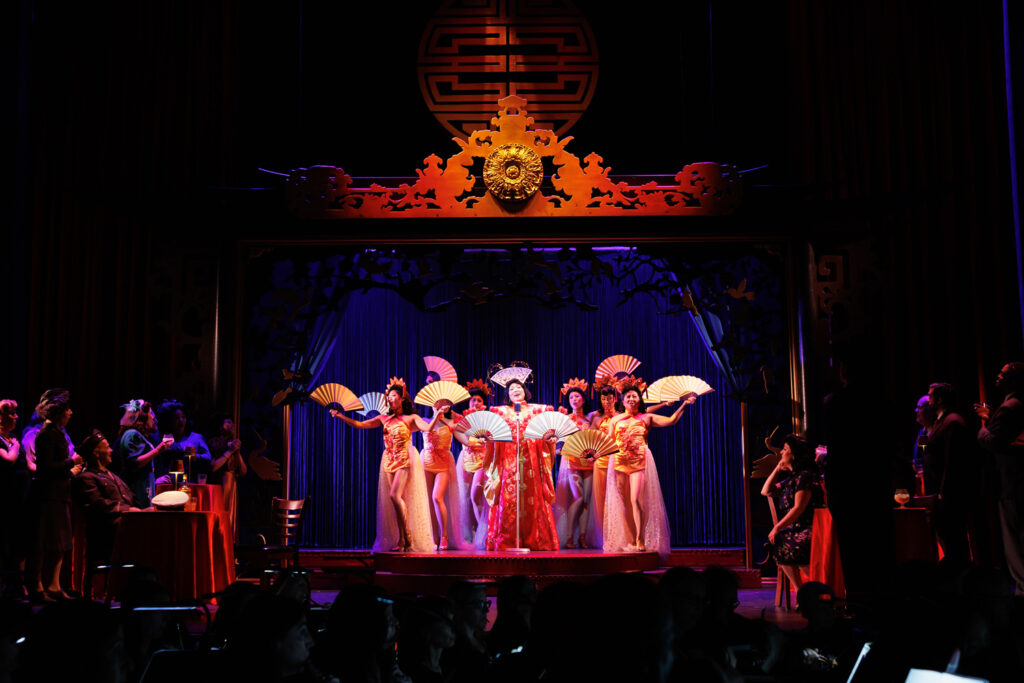An elaborate nightclub scene, in which patrons watch from the side as seven lavishly costumed entertainers with fans—including Butterfly, at a center mic—perform.