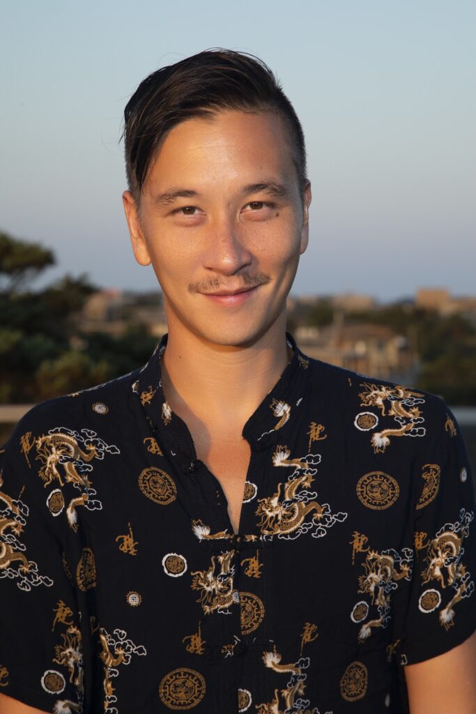 Chan, an Asian man with sort black hair wearing a black printed shirt, gives the camera a small smile.