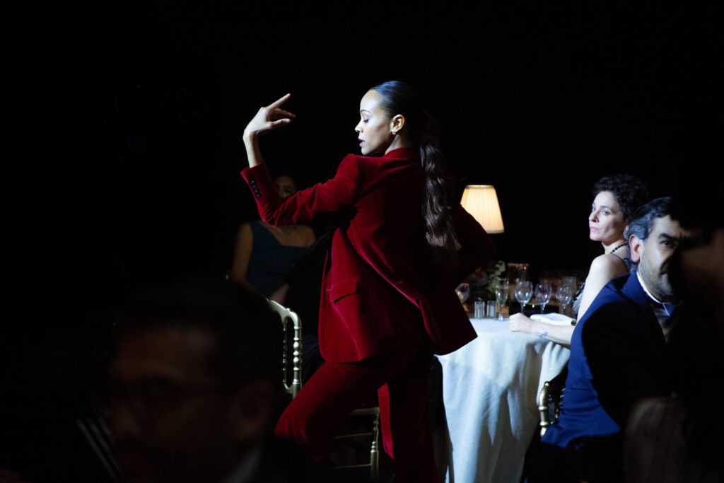 Saldaña—wearing a red velvet suit, her dark hair slicked back—stands in profile in a bright spotlight, her left arm raised and bent at the elbow toward her face, her fingers delicate. Behind her, glamorous diners sit at white-cloth-covered tables.