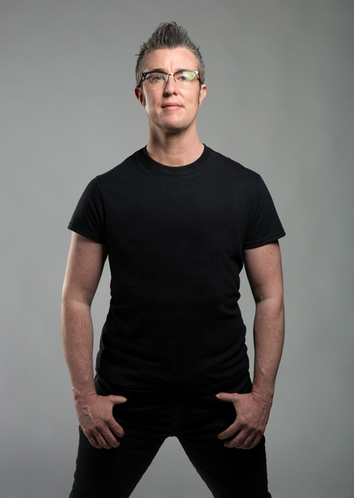 Dorsey—wearing a black t-shirt, black pants, and glasses, his short grey hair spiked into a fauxhawk—tucks his thumbs into his pockets as he looks directly into the camera, half smiling.