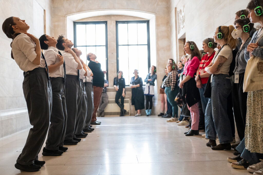 In a long windowed hallway, a group of performers stands in a line at the left, each leaning back slightly with a hand on their chest. A line of audience members wearing headphones stands opposite them, watching.